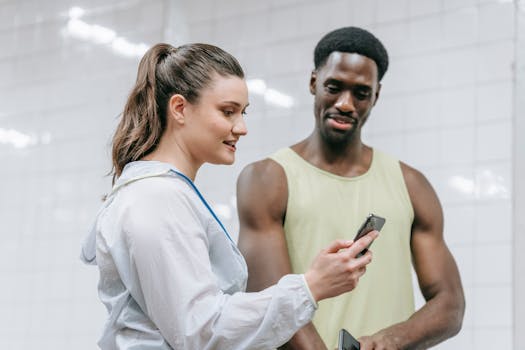 A black man and a caucasian woman using a smartphone in a gym setting, sharing workout insights.