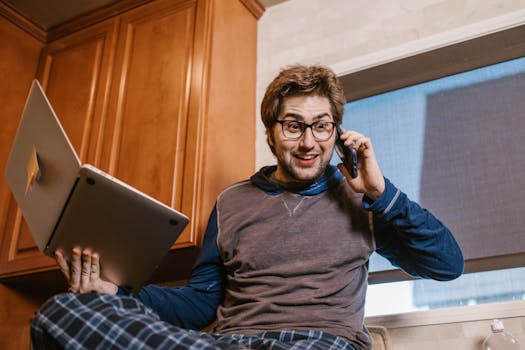 Caucasian man using laptop and phone for remote work at home, relaxed and multitasking.