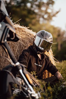 Motorcyclist in helmet sits by hay bale with motorcycle in sunny rural setting.